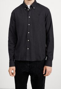 Camicia scura con bottoni, colletto, dotata di una trama sottile, bottoni bianchi e un piccolo logo sul petto. Materiale in cotone.
