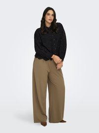 Vrouw draagt een zwarte blouse met lange mouwen en oogjes, losse bruine wijde broekspijpen en bruine puntige schoenen, staand tegen een effen lichte achtergrond.