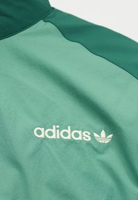 Πράσινο μπουφάν adidas με λευκό κεντημένο λογότυπο και σύμβολο τριφυλλιού στο στήθος, δείχνοντας την υφή του υφάσματος και τις ραφές.
