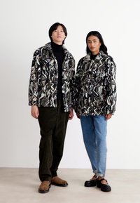 Twee personen staan naast elkaar, gekleed in jassen met abstracte zwart-, wit- en grijstinten. De jassen zijn pluche, gelaagd en losvallend.