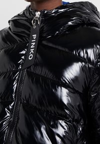 Zbliżenie na osobę w błyszczącej czarnej kurtce puchowej z kapturem i suwakiem z etykietą "PINKO".
