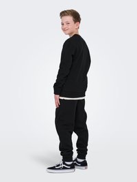 Un outfit composto da felpa nera e pantaloni della tuta neri su un ragazzo, caratterizzato da un taglio comodo, polsini a coste, abbinato a sneakers nere. T-shirt bianca visibile.