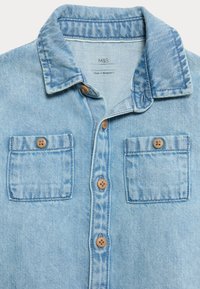 Camicia di denim azzurro chiaro con colletto, due tasche sul petto e bottoni marroni. Presenta una texture sbiadita e un design classico. Prodotta in Bangladesh.