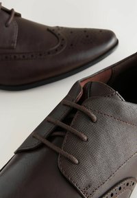 Chaussures habillées en cuir marron avec des perforations, sections lisses et texturées, et lacets plats. Semelle en caoutchouc noir.