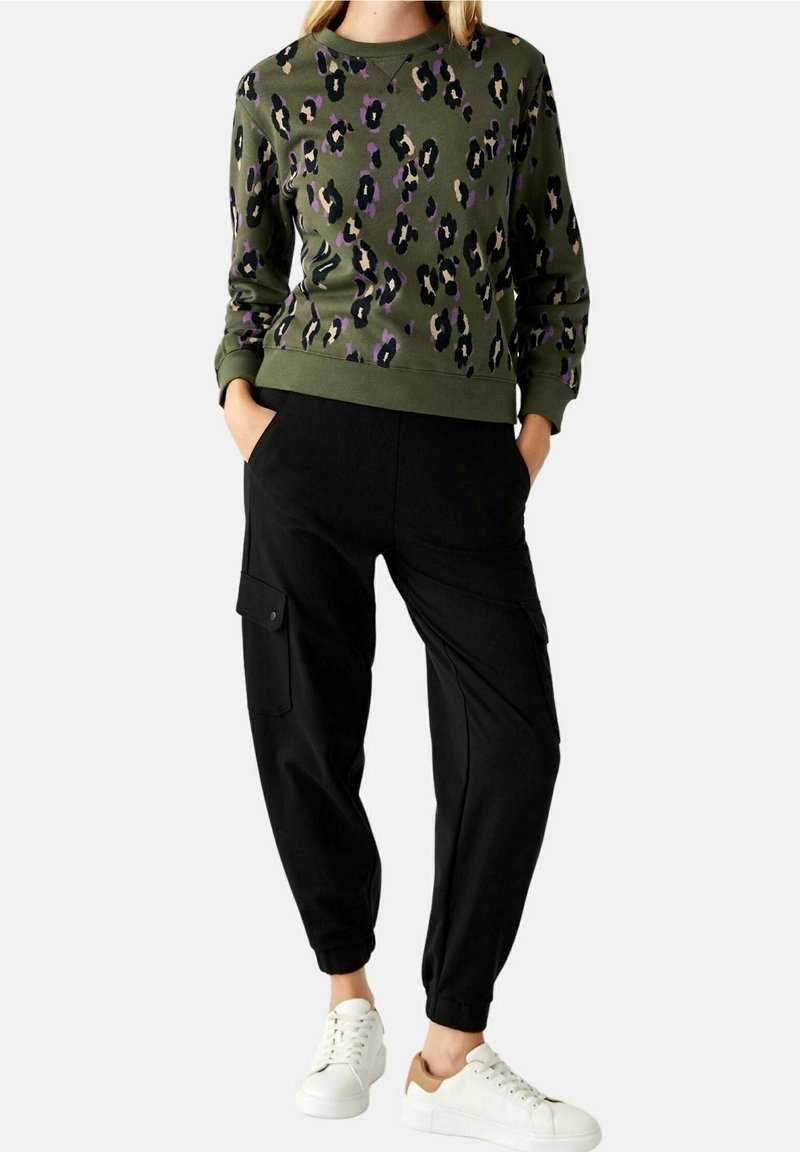 Sweatshirt vert avec imprimé léopard violet et noir, assorti à un pantalon cargo noir avec poches latérales et des baskets blanches.