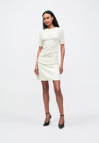 Egy göndör hajú nő áll, aki egy testhez simuló krémszínű, rövid ujjú felsőt és egy hozzá illő bordás szoknyát visel, fekete boka-örvös magassarkú cipőkkel párosítva.