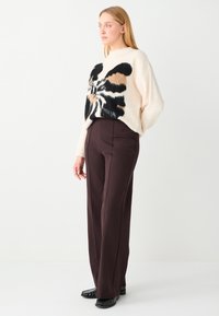 Oversized crèmekleurige trui met zwart en bruin abstract patroon, gecombineerd met hooggetailleerde bruine broek en zwarte schoenen, staande pose.