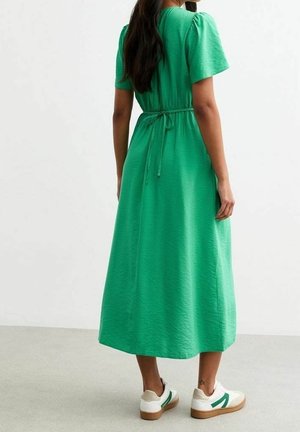 Vrouw die binnenshuis staat, gekleed in een groene midi-jurk met korte mouwen en witte sneakers met groene accenten, van de camera af gekeerd.