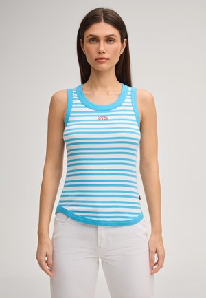 Frau trägt ein blau-weiß gestreiftes ärmelloses Oberteil mit "Joop! Jeans"-Logo, kombiniert mit weißer Hose, steht vor einem einfarbigen Hintergrund.