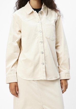 Vrouw draagt een crèmekleurig corduroy blouse met knopen en een borstzak, gecombineerd met een bijpassende rok, staand tegen een witte achtergrond.