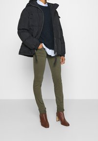 Svart puffjacka med huva, mörkblå tröja, lager av vit skjorta, olivgröna skinny jeans och bruna ankelstövlar med klossklack.