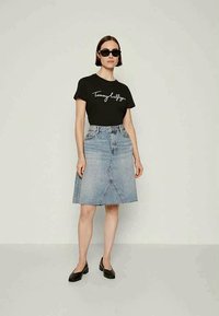 Schwarzes T-Shirt mit weißem Schriftlogo, kombiniert mit einem hellblauen Denim A-Linien-Rock mit rohem Saum und einem Schlitz vorne. Schwarze Ballerinas getragen.