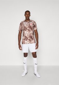 Brun kamuflasjemønstret fotballdrakt med logo, sammen med hvite shorts og knelange sokker. Modellen har på seg hvite joggesko.