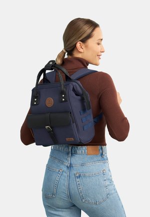 Marineblauer Rucksack mit schwarzen Akzenten, aus strapazierfähigem Material, ausgestattet mit Handgriffen oben, einem Vorderklappenverschluss mit Schnalle und verstellbaren Trägern.