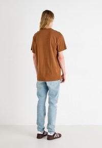 Camiseta de algodón marrón de manga corta, combinada con jeans de color azul claro. El modelo lleva zapatillas oscuras con rayas blancas, visto desde atrás.