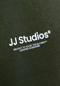 Tamnozeleni materijal s bijelim tekstom koji glasi "JJ Studios© Donosi vam J&J Crew™ Dizajnirano u Danskoj" u podebljanom i malom fontu.