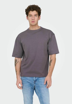 Jeune homme aux cheveux bouclés portant un T-shirt ample gris foncé et un jean bleu clair, debout avec une expression neutre sur un fond uni.