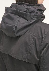 Svart lättviktsjacka med hög krage och huva, med en texturerad tyg och synliga sömsdetaljer på baksidan.