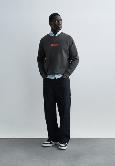 Άνδρας που στέκεται με τα χέρια στις τσέπες, φορά γκρι φούτερ με την επιγραφή "VOLTAIRE", γαλάζιο πουκάμισο, μαύρο παντελόνι και ασπρόμαυρα αθλητικά παπούτσια.
