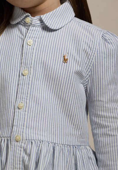 Barn iført en blå og hvid vertikalt striped skjorte med knapper og et lille broderet brunt hest og rytter logo på brystet.