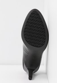 Zapato de tacón alto negro con parte superior de cuero liso, punta afilada y suela de goma con patrón. El tacón presenta una textura y un diseño únicos.