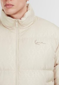 Gaiši bež pufota jakas ar augstu apkakli, ar smalku rakstu un priekšējo rāvējslēdzēju. Logo izšūts kontrastējošā krāsā.