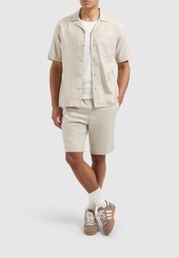 Lichtbeige kortärmige overhemd met knoopjes en een borstzak, gecombineerd met bijpassende shorts. Bruine sneakers met witte accenten en lichte sokken.