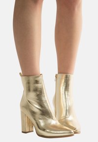 Golden metallic ankle boots with a pointed toe, high block heel, smooth texture, and side zipper detail.