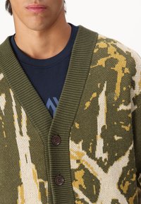 Grønn og kremfarget cardigan med knappelukking, V-halsdesign og strukturert strikkemønster med brune detaljer.