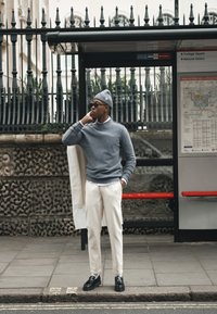 Mężczyzna w szarym swetrze, beżowych spodniach i czapce stoi na przystanku autobusowym, trzymając białą kurtkę, spoglądając w bok w pobliżu wskazówek do Trafalgar Square.