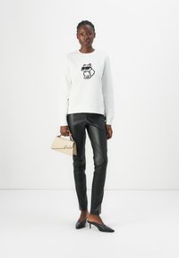 Vit sweatshirt med en broderad rävdesign, i kombination med svarta läderbyxor. Modellen håller en beige handväska och bär svarta klackar.