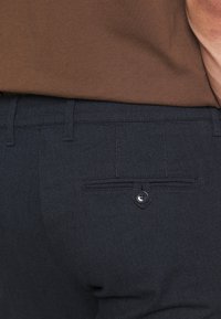 Pantalons bleu foncé en tissu texturé. Dotés d'une poche arrière avec une fermeture à bouton et de passants de ceinture. Associés à une chemise marron.