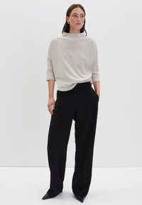 Frau steht da, trägt ein hellbeiges, strukturiertes Oberteil mit Ellenbogenärmeln und lockeren, schwarzen, weiten Hosen, die Hände teilweise in den Taschen.