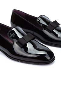 Mocasines de charol negro con punta redondeada, que presentan un lazo de cinta negro en el empeine, con un forro interior púrpura.