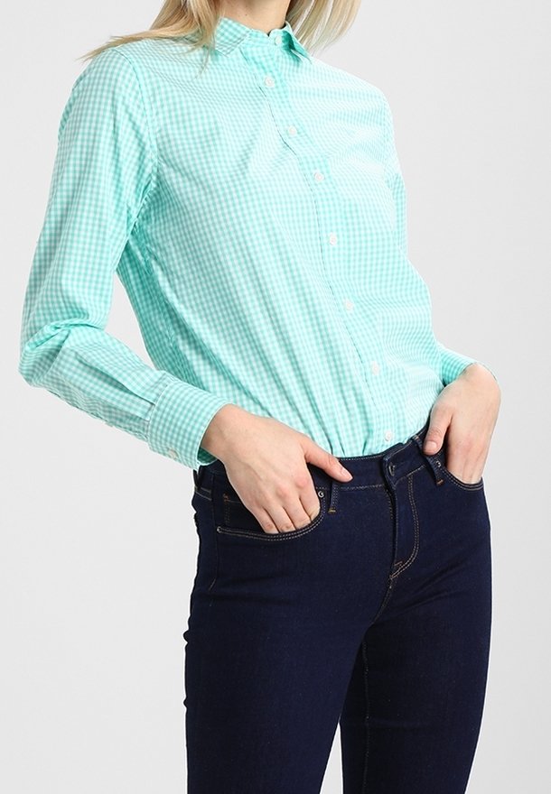 Femme portant une chemise à carreaux vert clair à manches longues, rentrée dans un jean bleu foncé, debout les mains dans les poches.