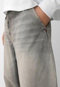 Harmaat farkut haalistuneella pinnalla, oranssilla ompeleilla, sivutaskulla ja rentoa istuvuutta tarjoavalla muotoilulla.