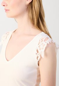 Krémový tílko s jemně vroubkovanou strukturou, zdobené bílou krajkovou květinovou aplikací na ramenech a hlubokým výstřihem do V.