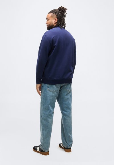 Άνδρας με ντραντλοκς που φοράει ναυτικό μπλε φούτερ, ανοιχτό μπλε τζιν και μαύρα-άσπρα αθλητικά παπούτσια, στέκεται και κοιτάζει προς τα αριστερά σε λευκό φόντο.