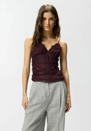 Donna con lunghi capelli castani che indossa una canottiera di pizzo viola scuro e pantaloni a vita alta grigio chiaro plissettati, in piedi davanti a uno sfondo neutro.