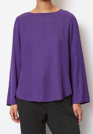 Femme portant un chemisier violet à manches longues avec un col rond et un pantalon noir, debout devant un fond blanc.