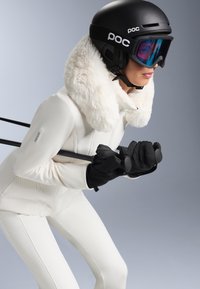 Costum de schi alb cu guler din blană sintetică, mănuși negre și cască. Schior ținând bețele, cu ochelari de schi cu nuanță albastră ce reflectă lumina. Textură netedă.