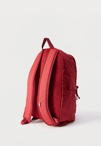 Κόκκινο σακίδιο πλάτης με επενδεδυμένους ιμάντες ώμου, άνω χειρολαβή και πλαϊνή τσέπη με φερμουάρ, όρθιο σε λευκό φόντο.