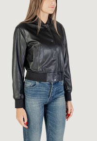 Giubbotto bomber in ecopelle nera con colletto, bottoni a pressione e due tasche laterali. Polsini e vita in costina. Indossato con jeans in denim blu.
