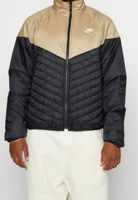 Chaqueta acolchada con diseño de bloques de color en negro y marrón claro, con cuello alto, cierre frontal con cremallera y textura acolchada en las secciones negras.