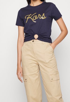 Námořnické tričko s kruhovým uzlovým detailem v pase, s kovovým zlatým nápisem „Kors“. Kombinováno s béžovými cargo kalhotami.