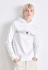 Hvit hettegenser med logo foran som leser "GANT 1949 AMERICAN SPORTSWEAR," med avslappet passform og ribbestrikkede mansjetter.