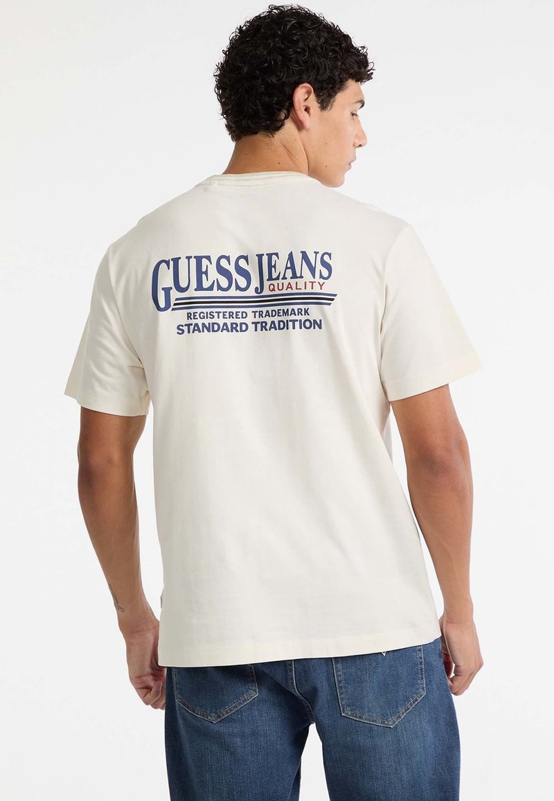 Młody mężczyzna z kręconymi ciemnymi włosami, ubrany w białą koszulkę z napisem "Guess Jeans Quality" i niebieskie dżinsy, pokazany od tyłu na białym tle.