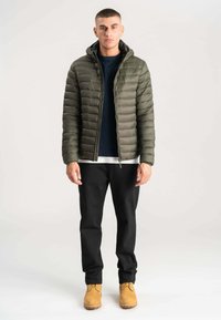 Oliven grøn pufferjakke med lynlås foran, hætte og quiltet design; parret med en navy sweater, sorte bukser og brune støvler.