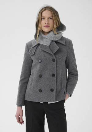 Femme portant un manteau gris croisé avec une capuche en maille et une écharpe, debout avec une main dans la poche devant un fond uni.