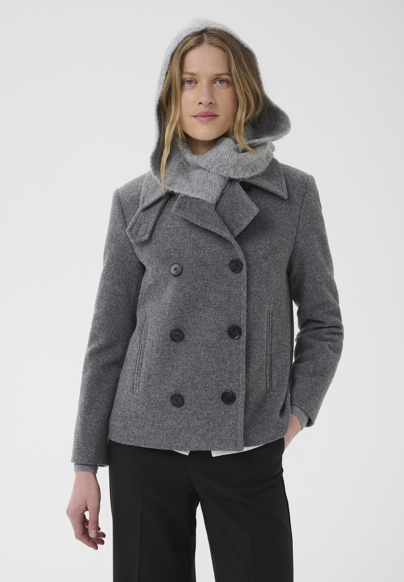 Femme portant un manteau gris croisé avec une capuche en maille et une écharpe, debout avec une main dans la poche devant un fond uni.